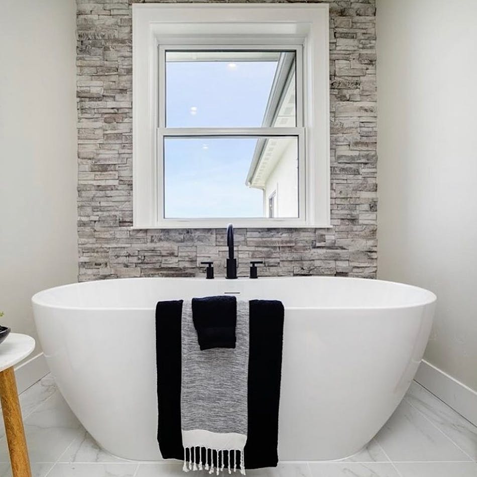 This bathroom design features a white bathtub, black fixtures and white stool and linens hanging on the side of the tub.