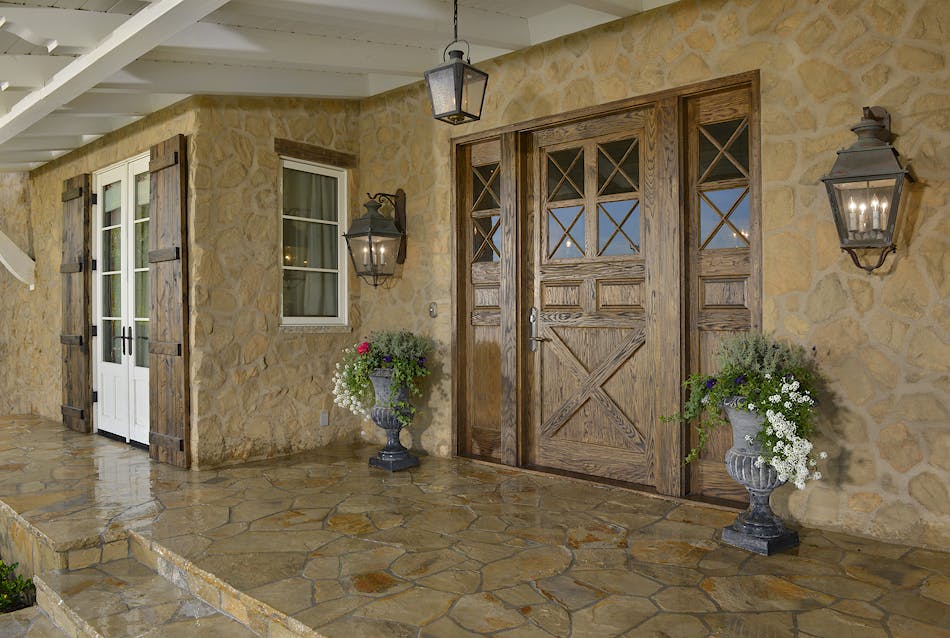 The front door of a Tuscan-style home. The home has a wooden door, two sconces, and stone flooring.