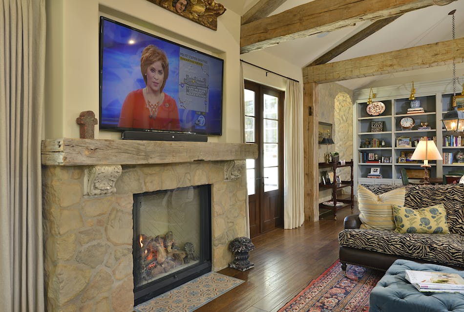 A living room with an exposed stone fireplace and exposed wood beams. There is a TV mounted above the fireplace and a zebra-patterned couch.