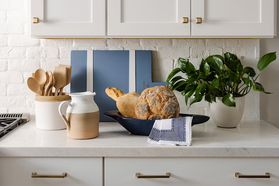 This kitchen features a white brick veneer backsplash. The counter has loaves of bread, a plant, a pitcher, a ceramic container holding wooden utensils and a blue cutting board on it.