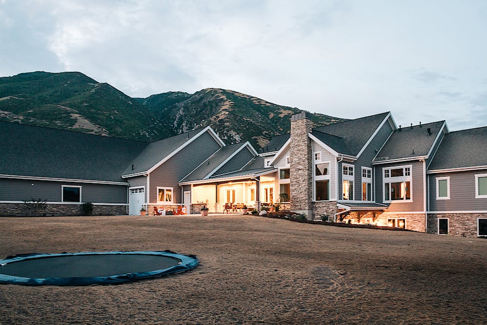 The backyard of a house at dusk with outdoor lights illuminating the back of the house. The backyard has an inground trampoline and behind the house is a mountainscape.