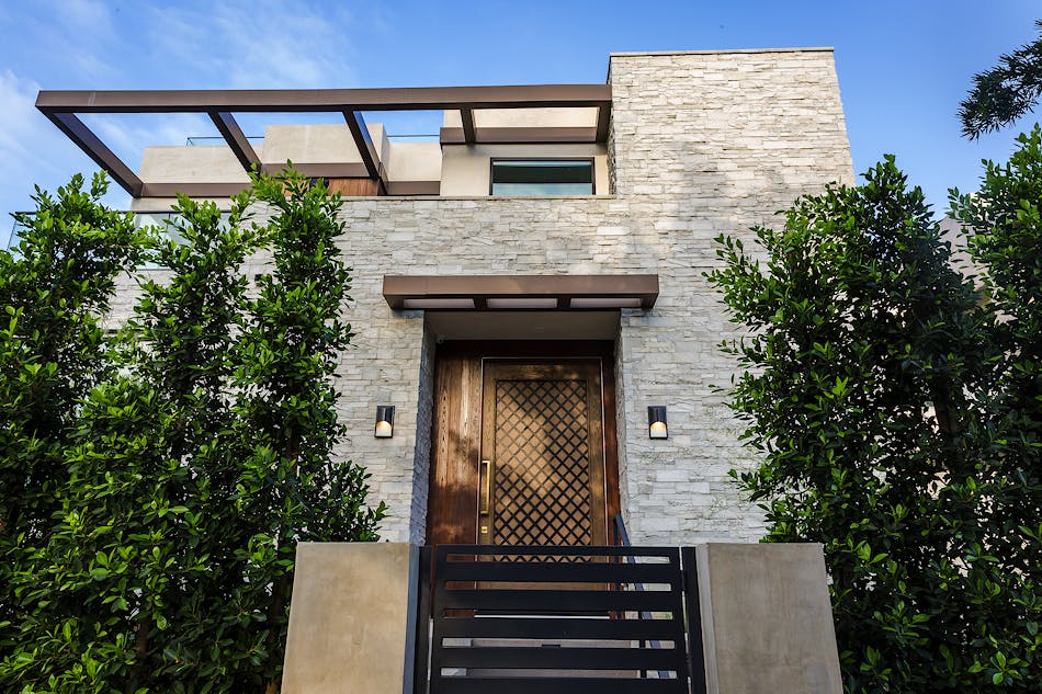 A modern home with light gray stone veneer siding and a unique wooden front door. The home has a metal gate with large trees on either side of the gate.