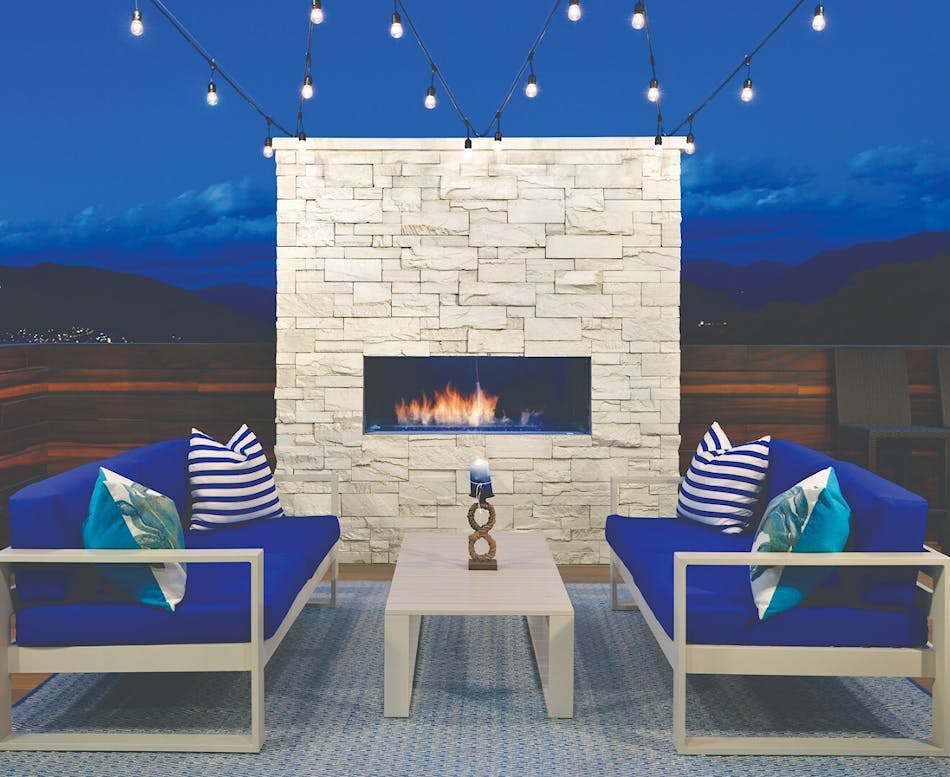 An outdoor fireplace at dusk with beige stone veneer, two blue couches, and a white coffee table.
