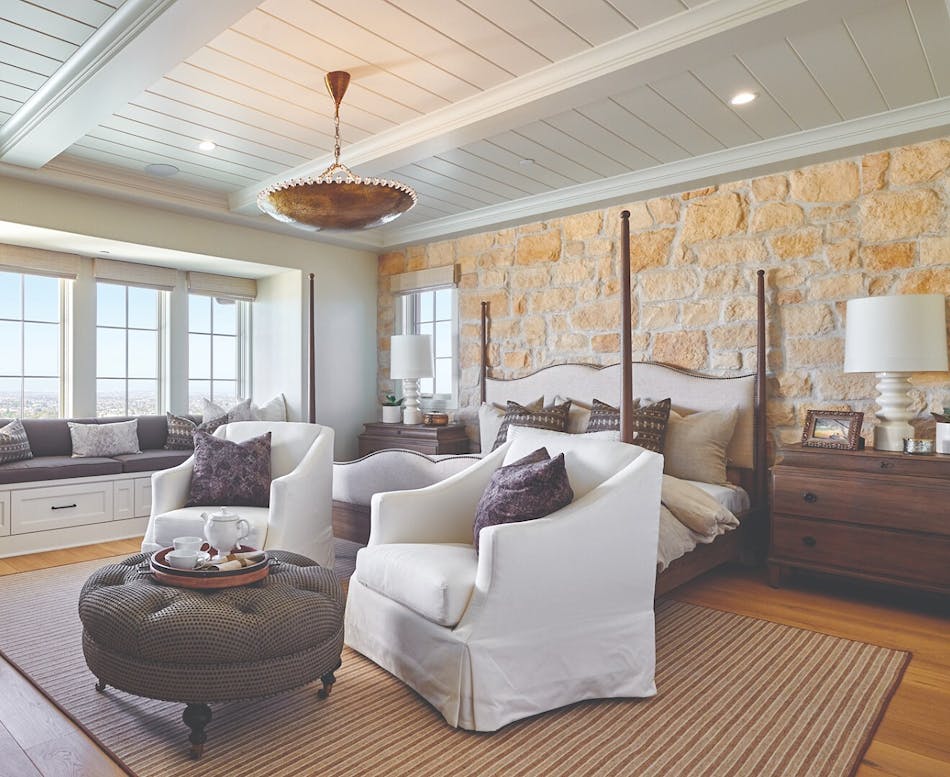 Master bedroom with white walls, four post bed, two white chairs in front of a round coffee table, and a masonry veneer accent wall behind the bed.