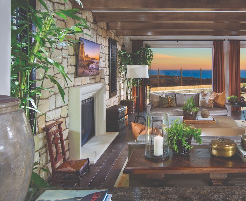 An accent wall of stone veneer houses a fireplace and looks out on an open room with dark wooden furniture, green potted plants, and a view out beyond the ocean sunset.